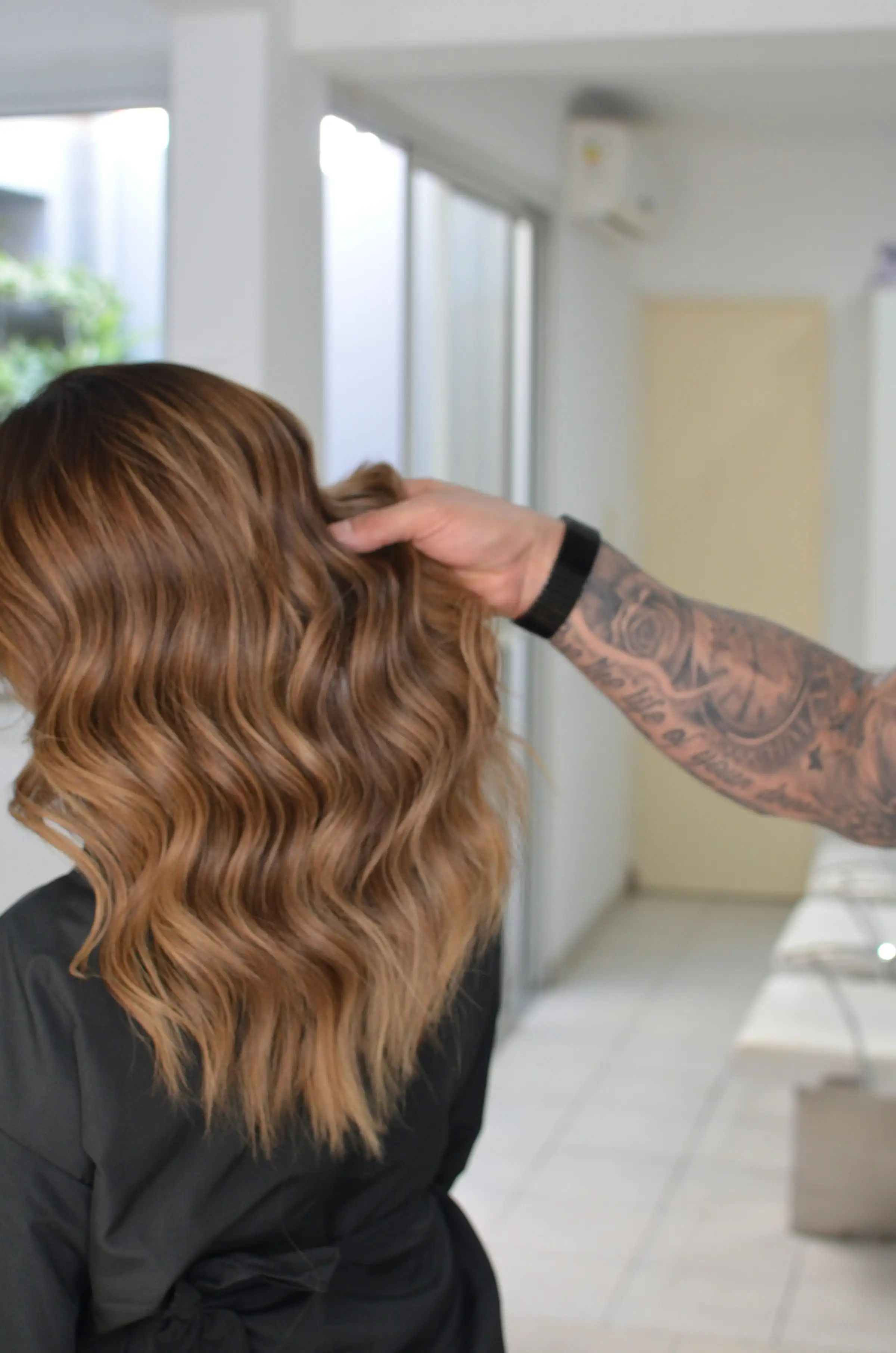A tattooed hand lifting long caramel balayage waves in a bright salon