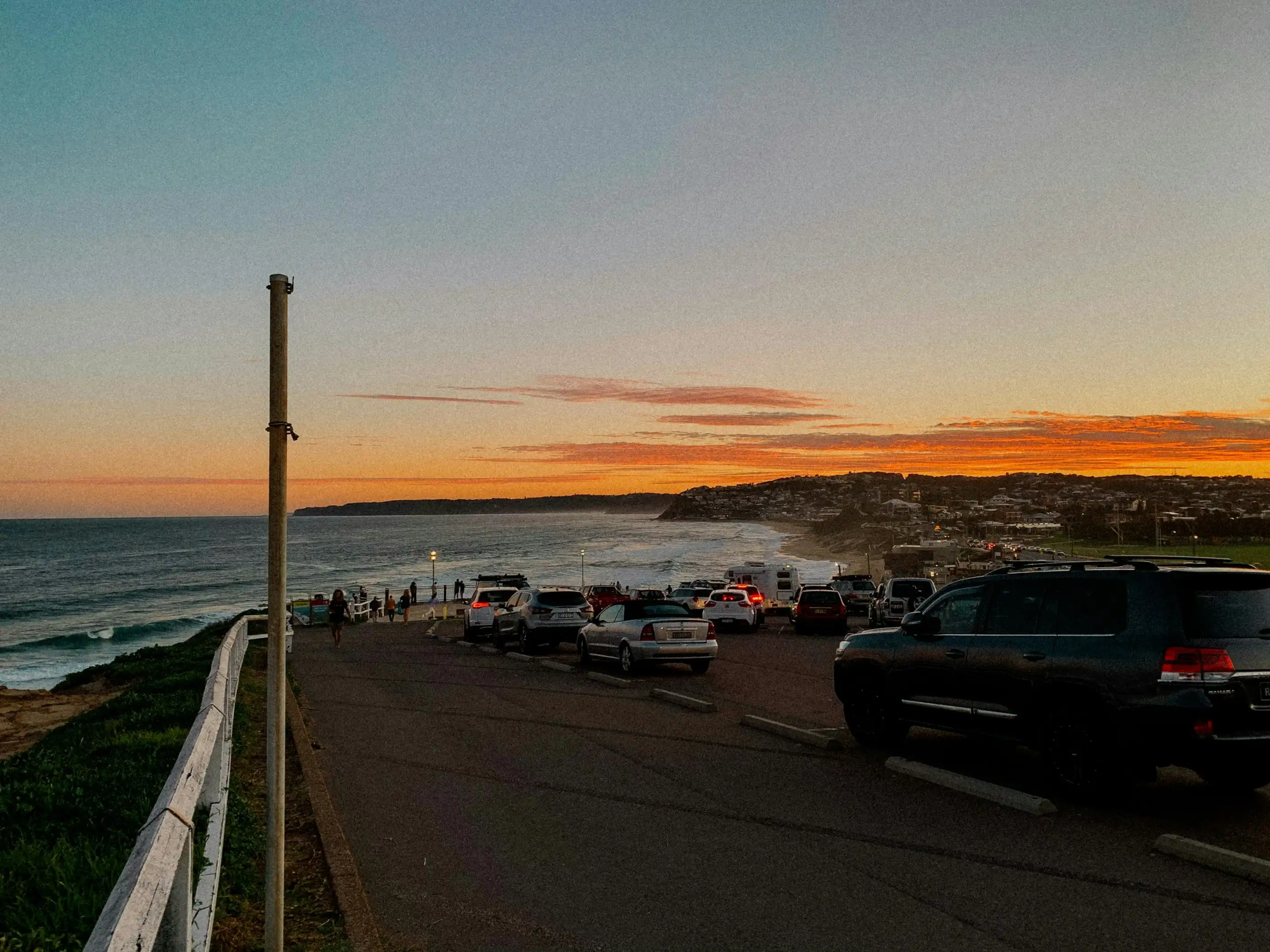 Cars parked along the clifftop road above Bar Beach at sunset, the coast curving away to the south