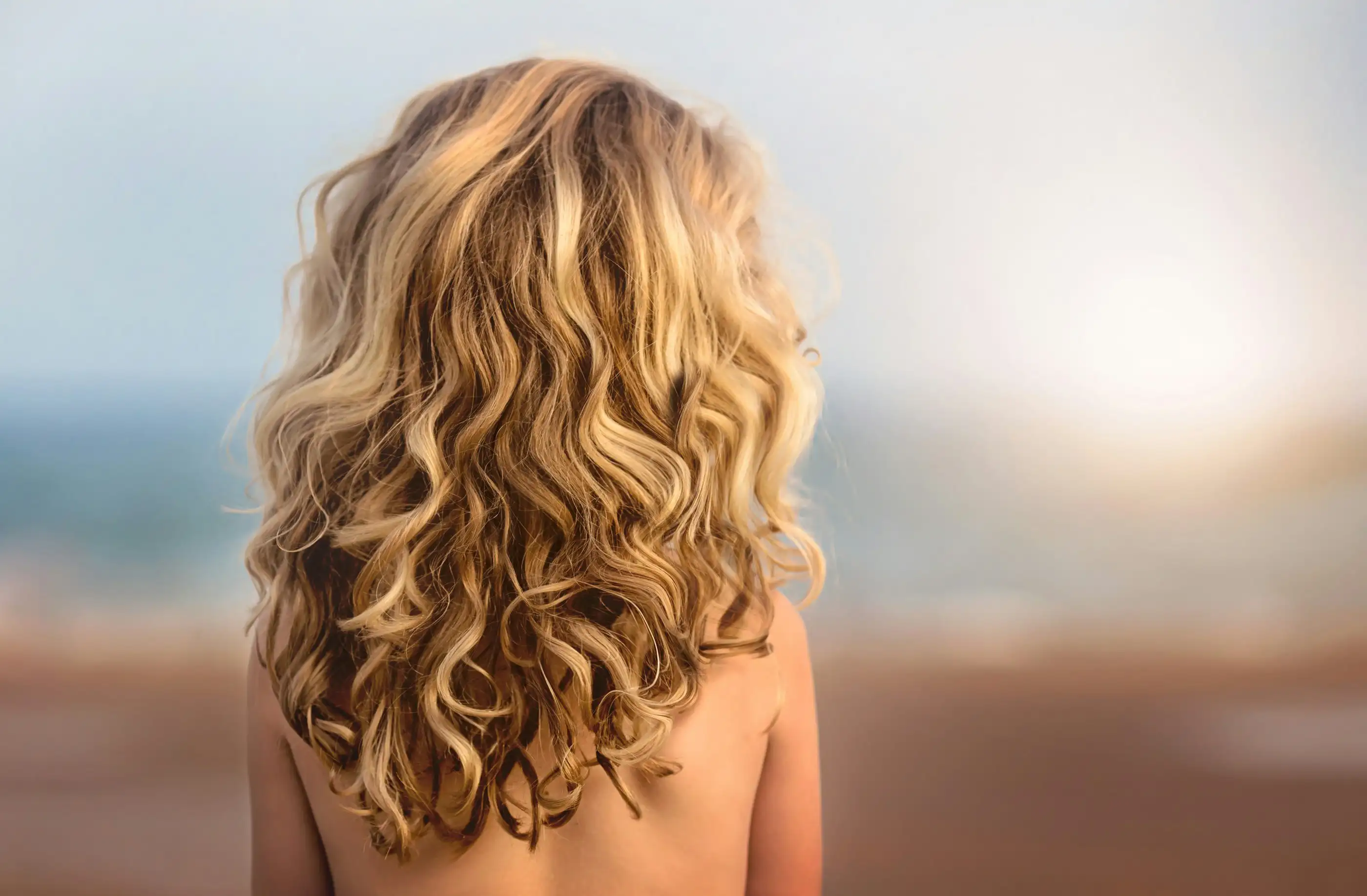 Long blonde curls seen from behind, with a blurred beach in the background