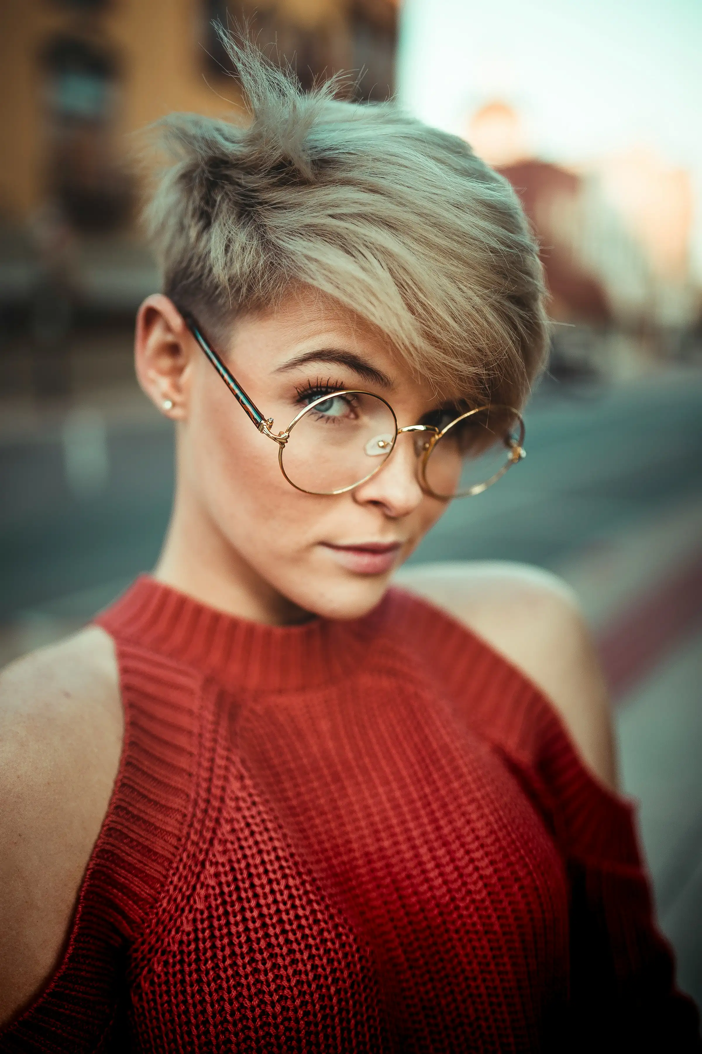 A short blonde pixie cut with round glasses and a red knit top
