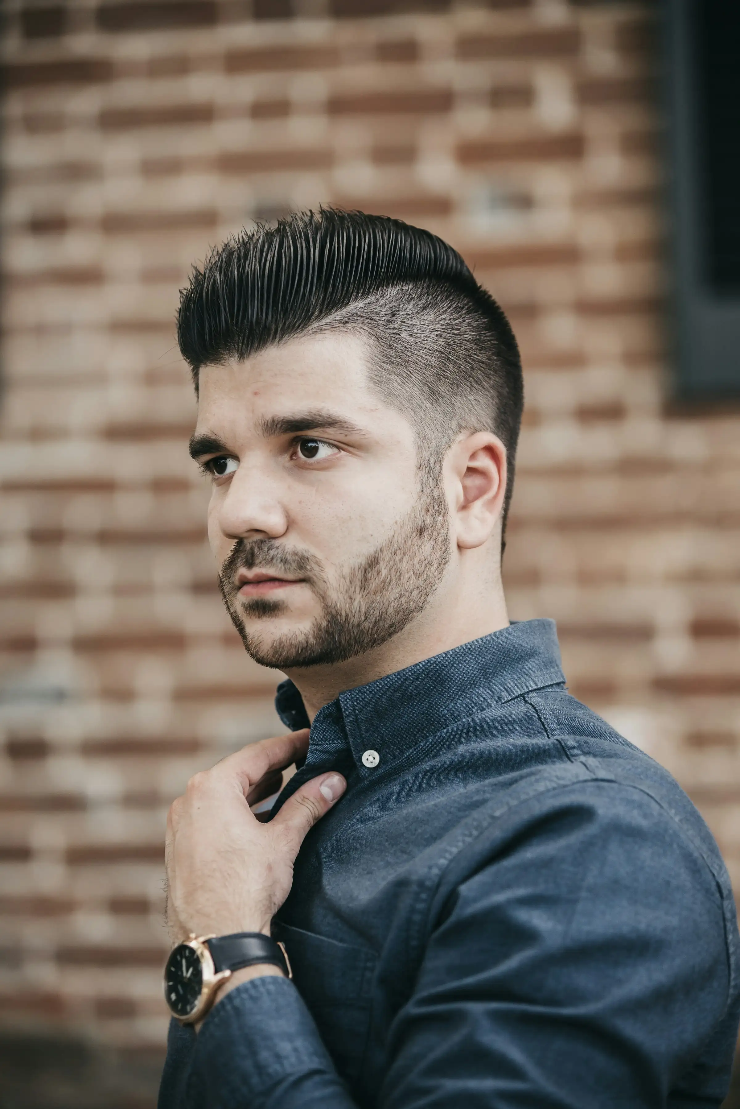 A man with a high, textured quiff and short faded sides, against a brick wall