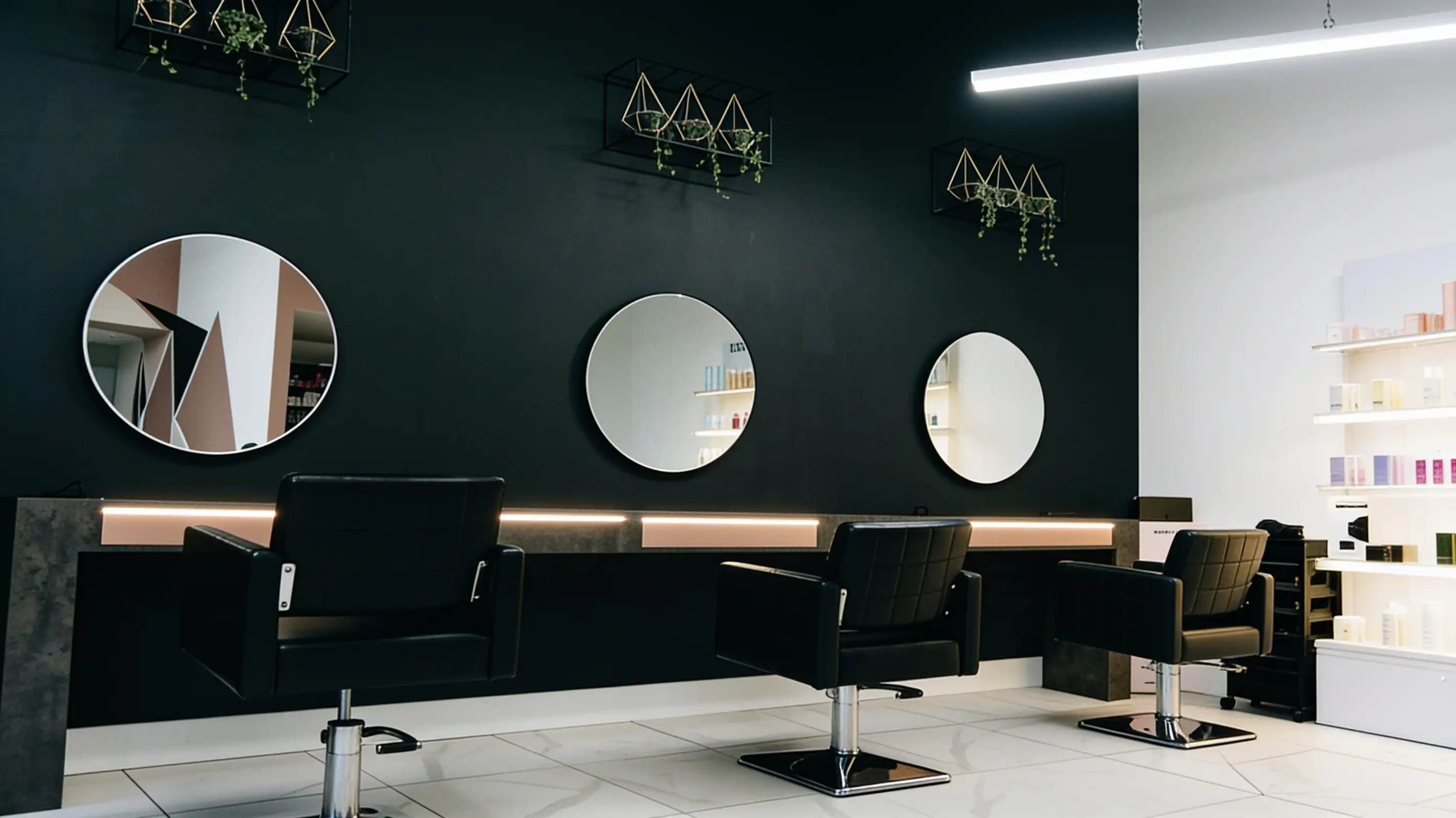 An empty salon seen at an angle: black styling chairs, round mirrors on a dark wall and white product shelves