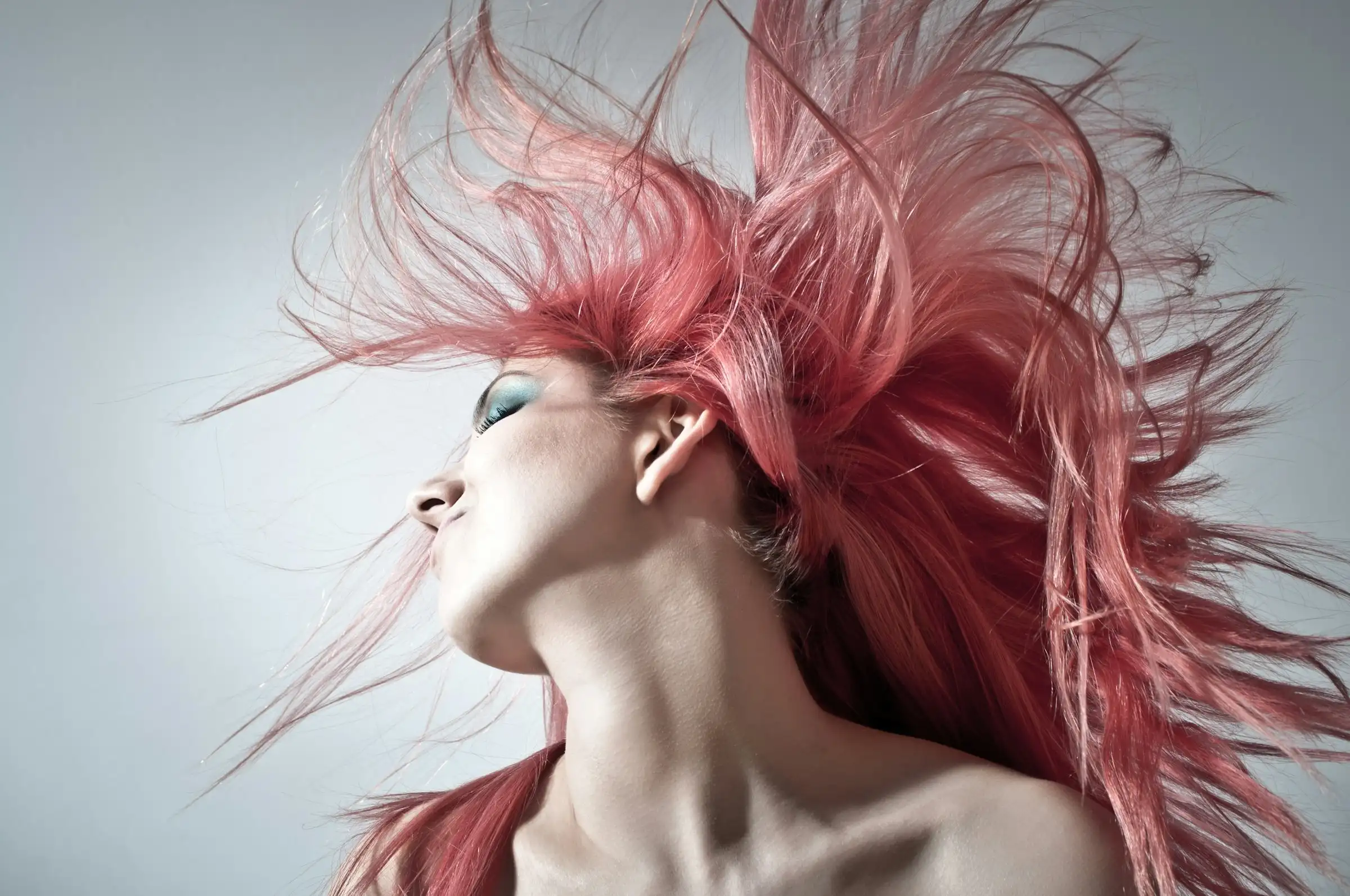 A woman throwing her head back, long pink hair flying out behind her against a pale grey backdrop